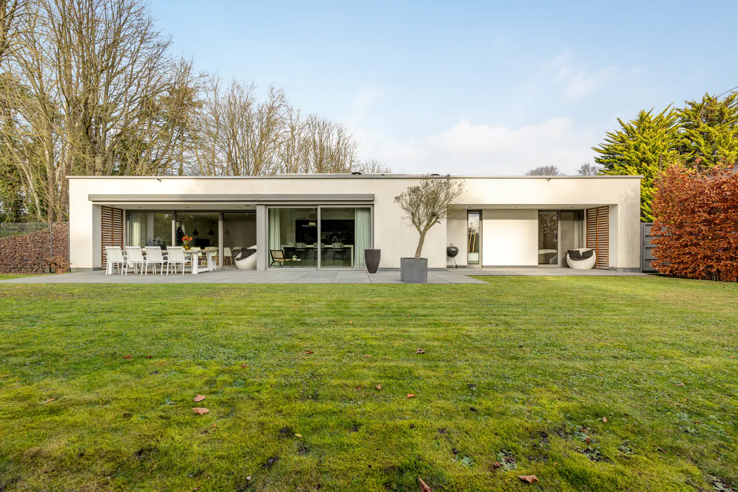 Modern, single-story white house with a flat roof, large windows, and a green lawn. Patio with dining set and lounge chairs. Trees in background.