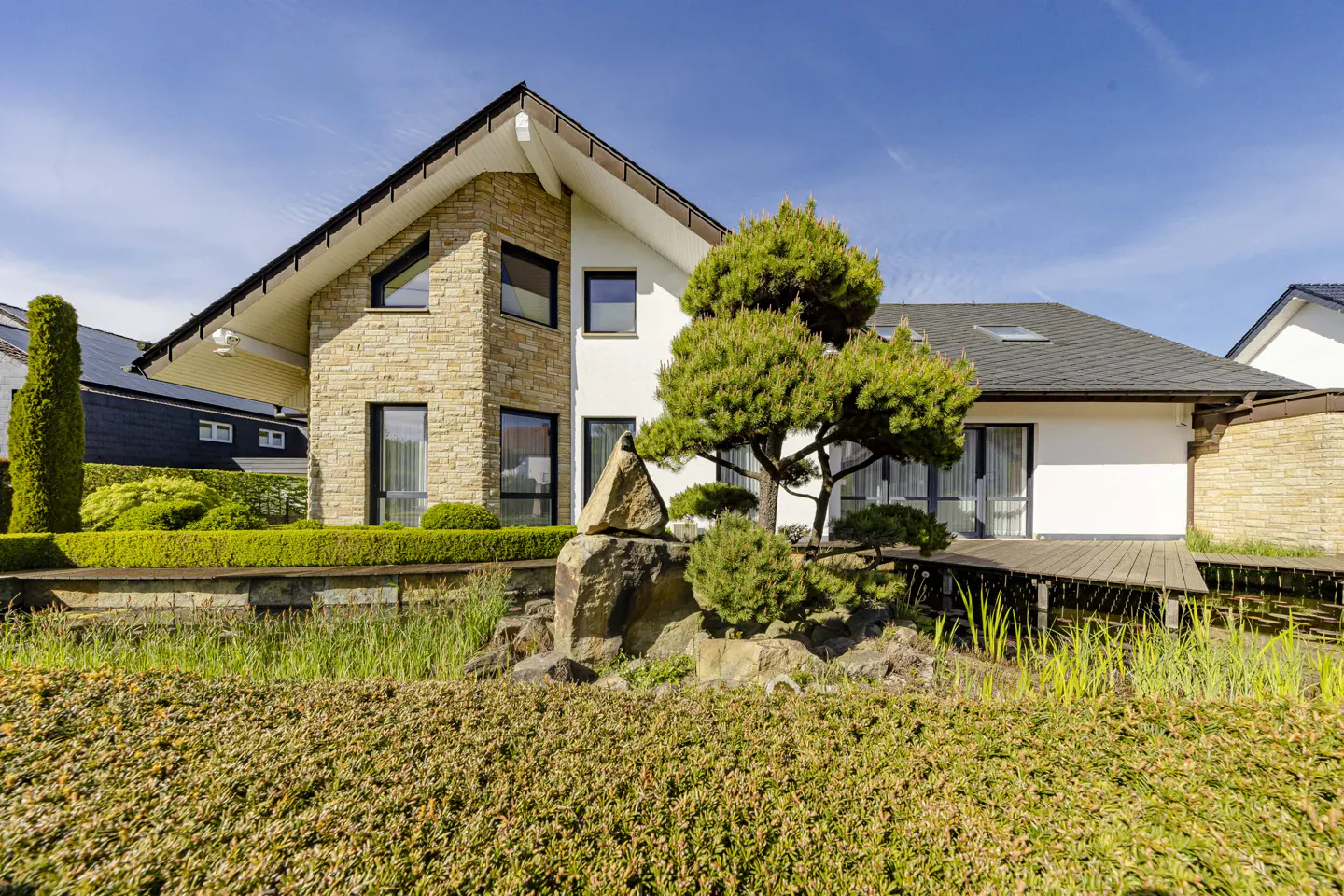A modern house with stone and white siding, black framed windows, and a landscaped yard with a pond and wooden bridge.