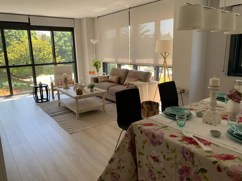 Bright living and dining area with large windows. A beige sofa, white coffee table, and floral tablecloth add charm to the space.