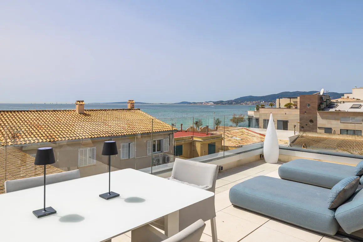 Rooftop patio with white table, chairs, and blue lounge chairs overlooking the ocean and city buildings on a sunny day.