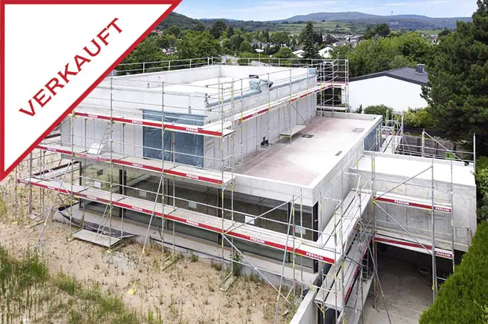 A modern, concrete house under construction is surrounded by scaffolding. A red triangle in the corner reads "VERKAUFT."