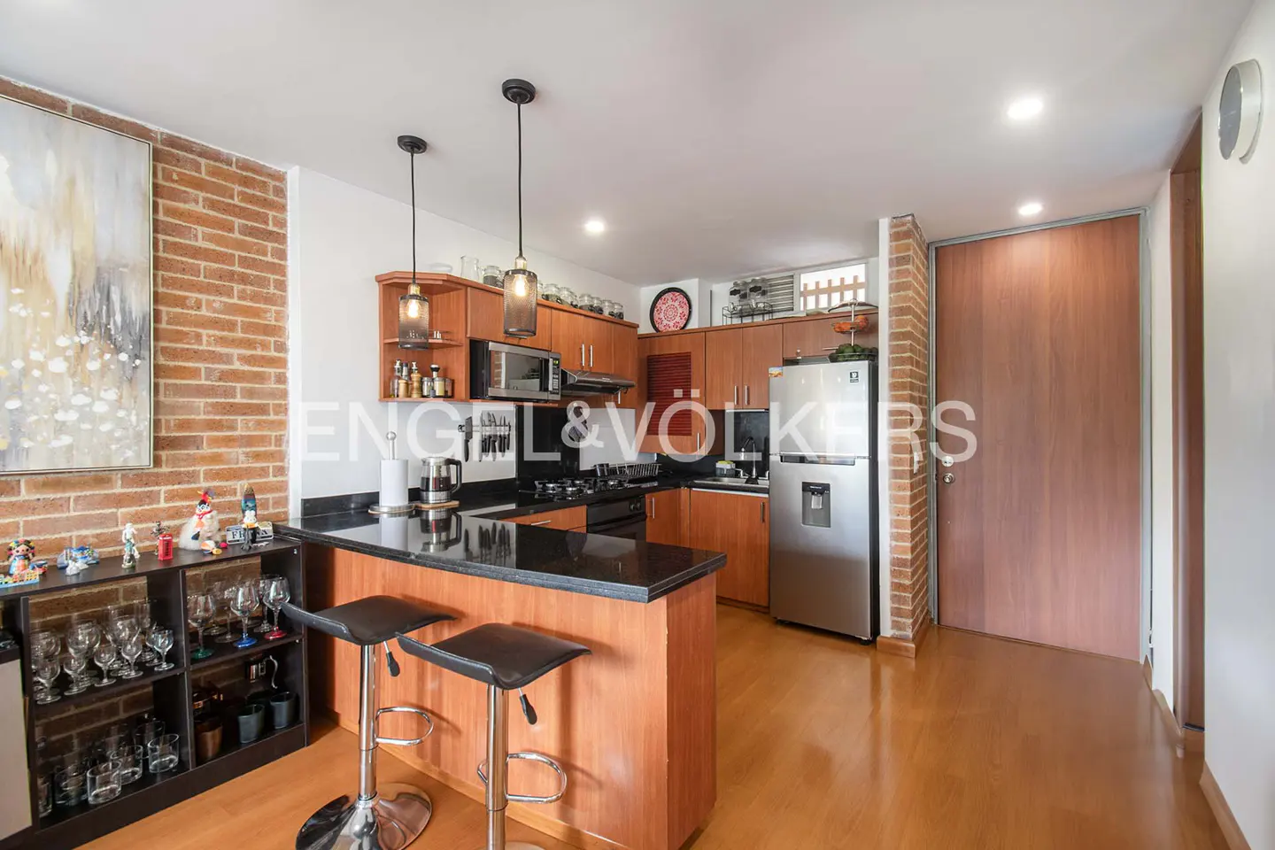 A kitchen with wood cabinets, black countertops, and stainless steel appliances. Two black stools sit at the island. A brick wall and wood floor add warmth.