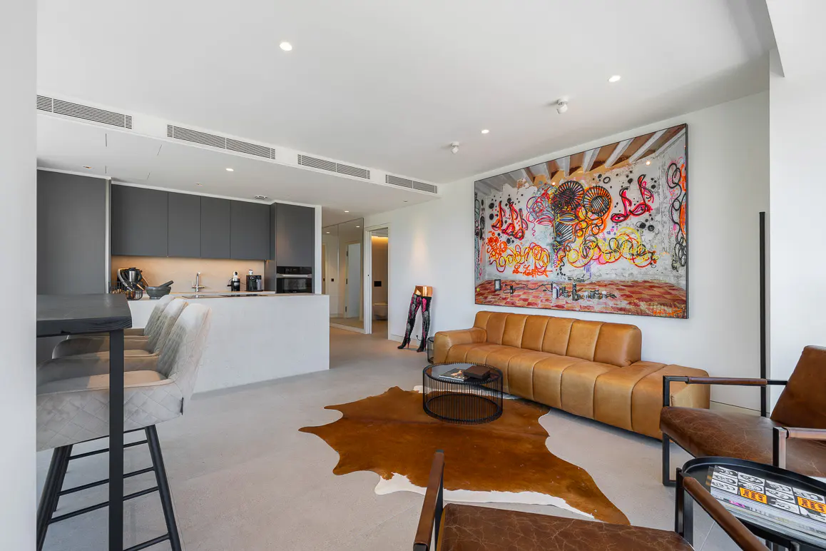 Modern open-plan living area with a tan leather sofa, cowhide rug, and large abstract graffiti art on a white wall.