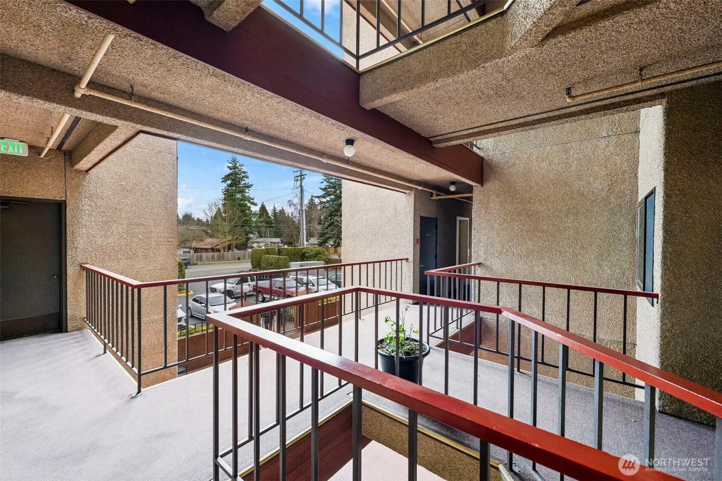 Outdoor apartment walkway with textured beige walls, red metal railings, and a view of a parking lot and trees.