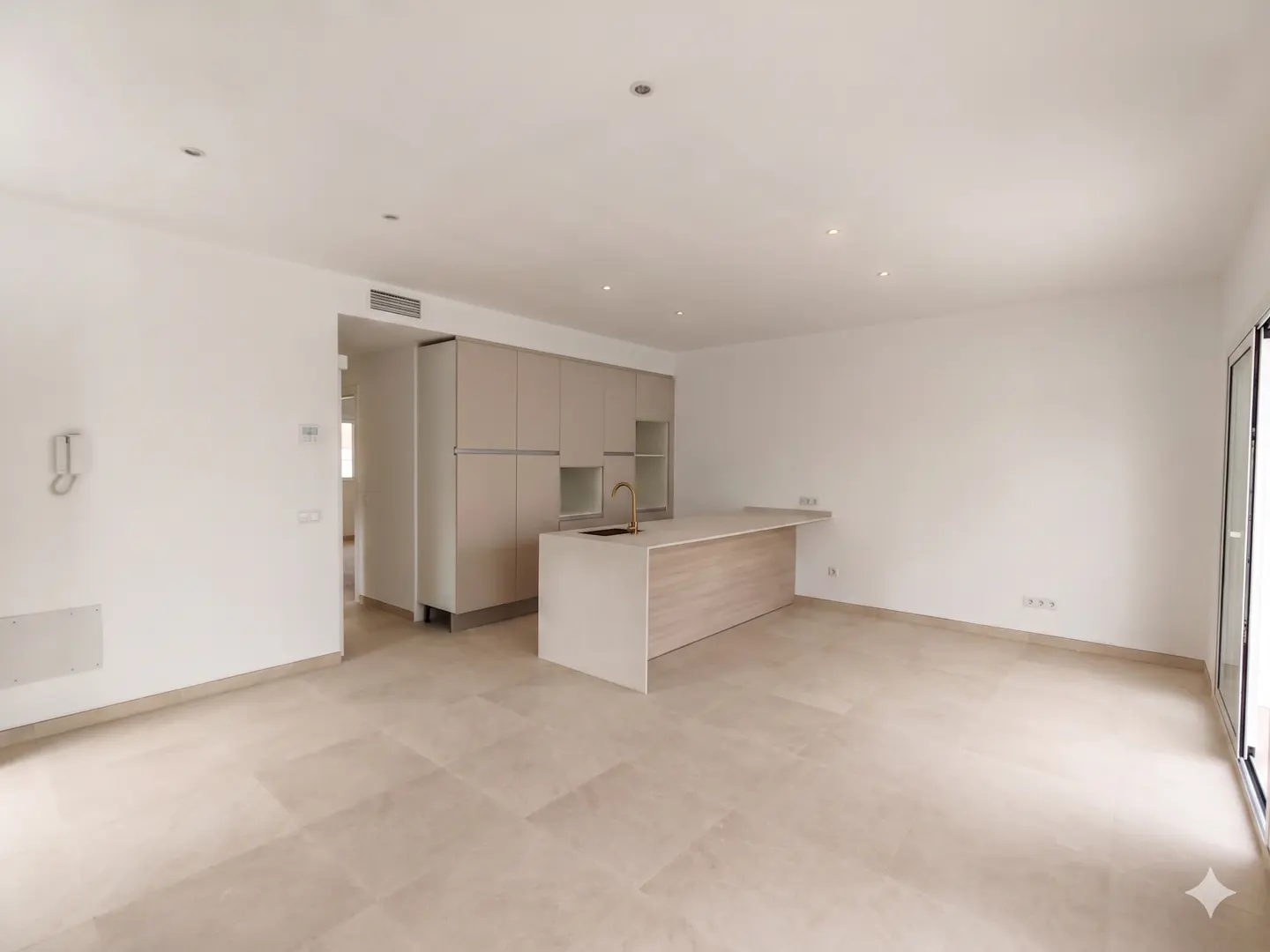 Bright, open-plan kitchen and living area with beige cabinets, island with gold faucet, and light tile flooring. White walls and recessed lighting.