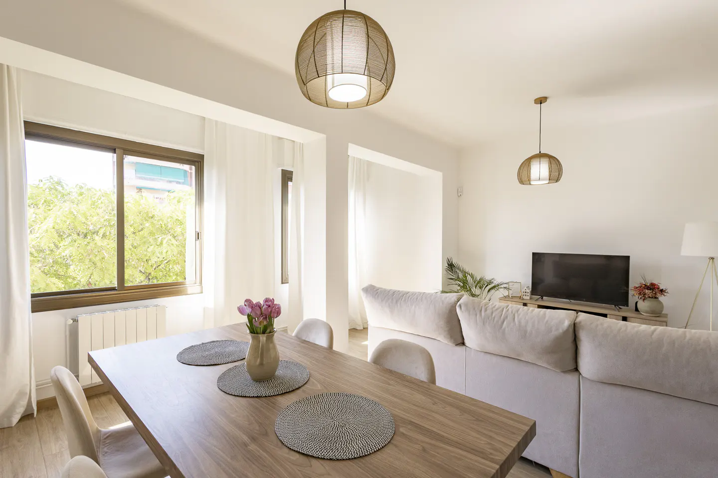 Bright, modern living room with a wood dining table, beige sofa, and woven pendant lights. Large window with green trees outside.