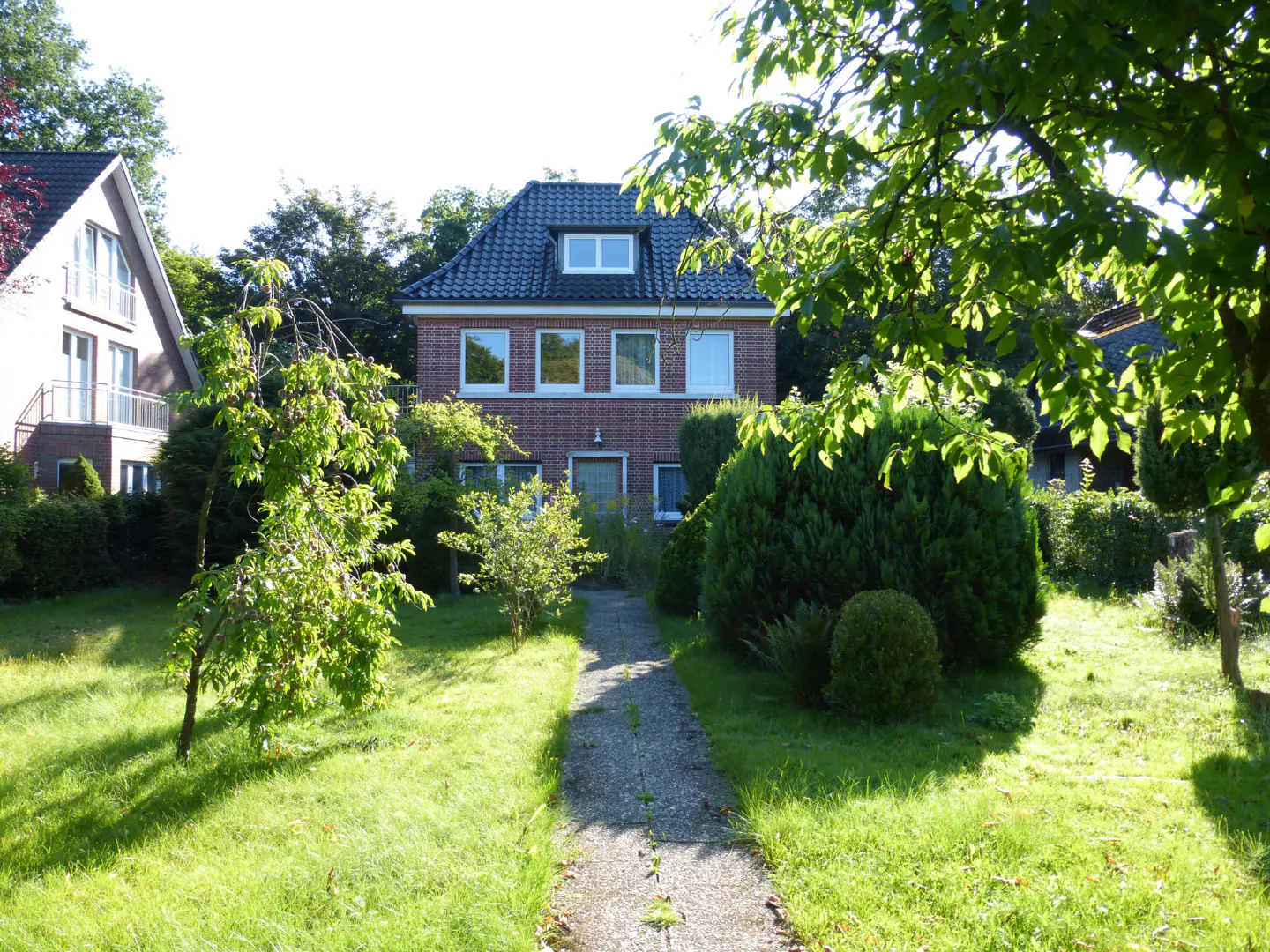 A stone path leads through a green lawn to a two-story red brick house with a dark tiled roof and lush garden surroundings.