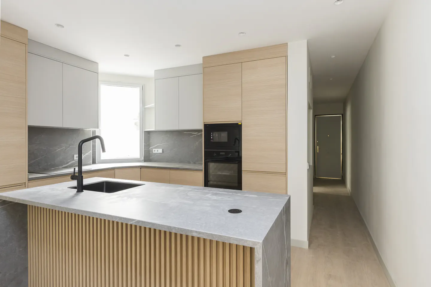 A modern kitchen with light wood and gray cabinets, a gray countertop island with a black faucet, and a hallway leading to a door.