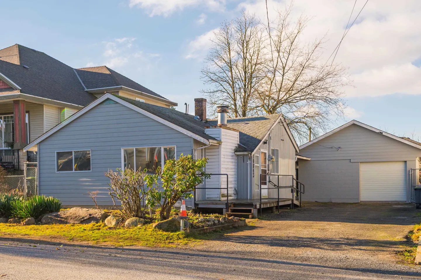 A light blue single-story house with a detached garage, a small front porch, and a gravel driveway on a sunny day.