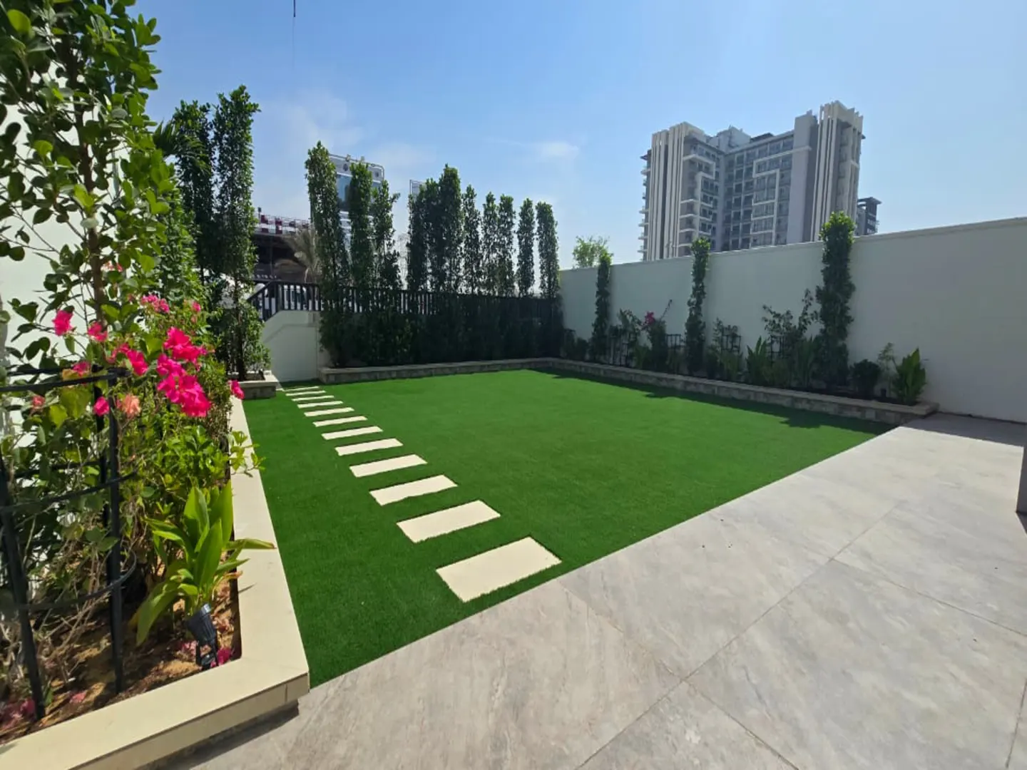 Backyard with artificial turf, stone stepping stones, a flower bed with pink flowers, and a tall apartment building behind.