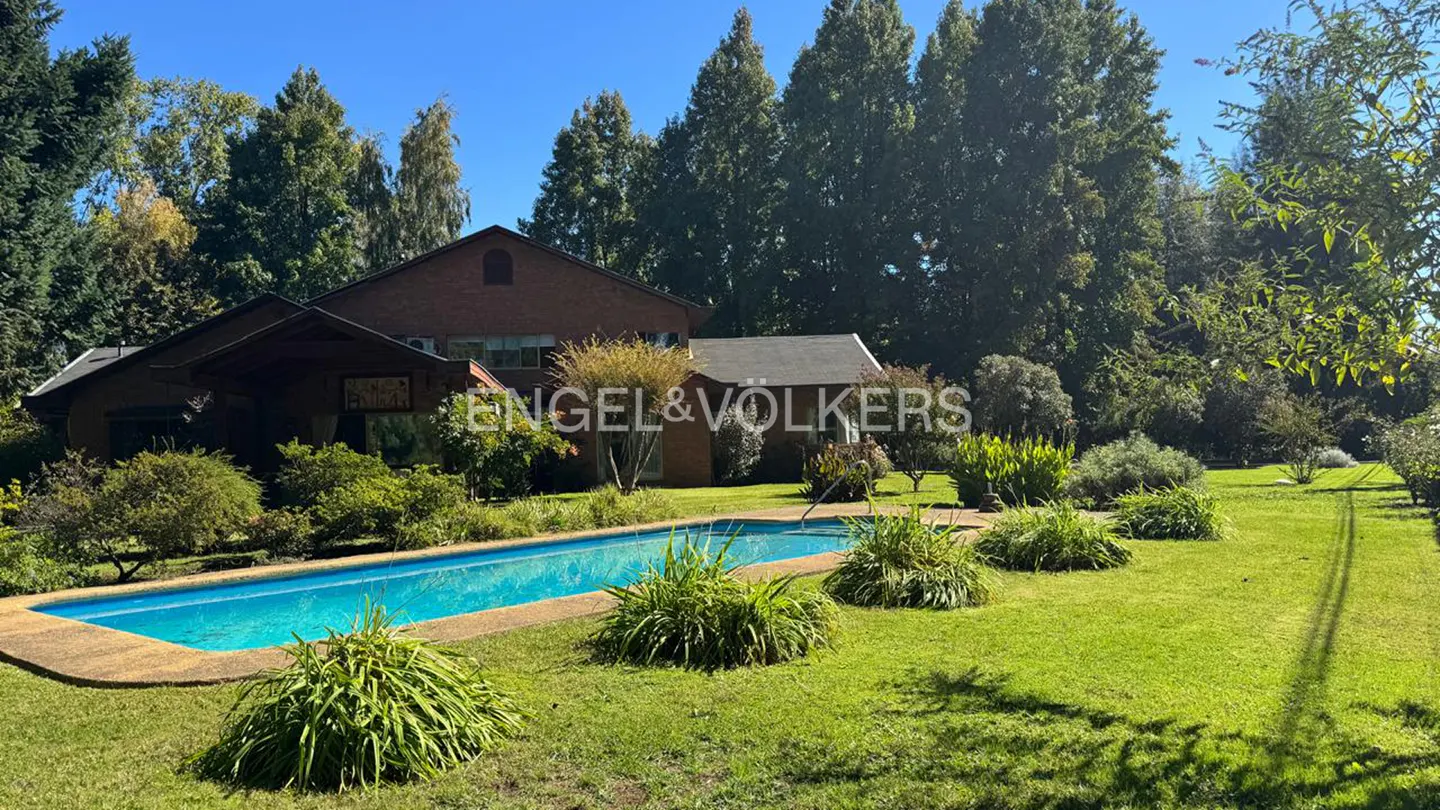Exterior of a brick house with a pool and green lawn, under a blue sky. Engel & Völkers logo is visible.