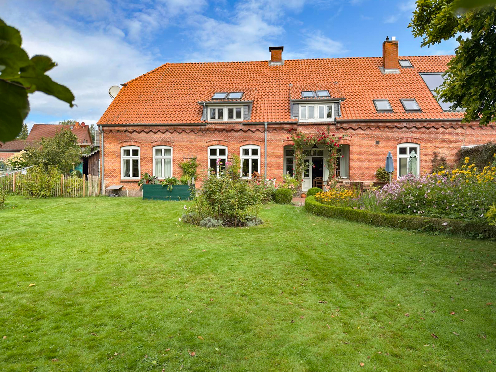 A red brick house with an orange tile roof, white windows, and a large, lush green lawn with flower beds and a garden. [AI-generated text]