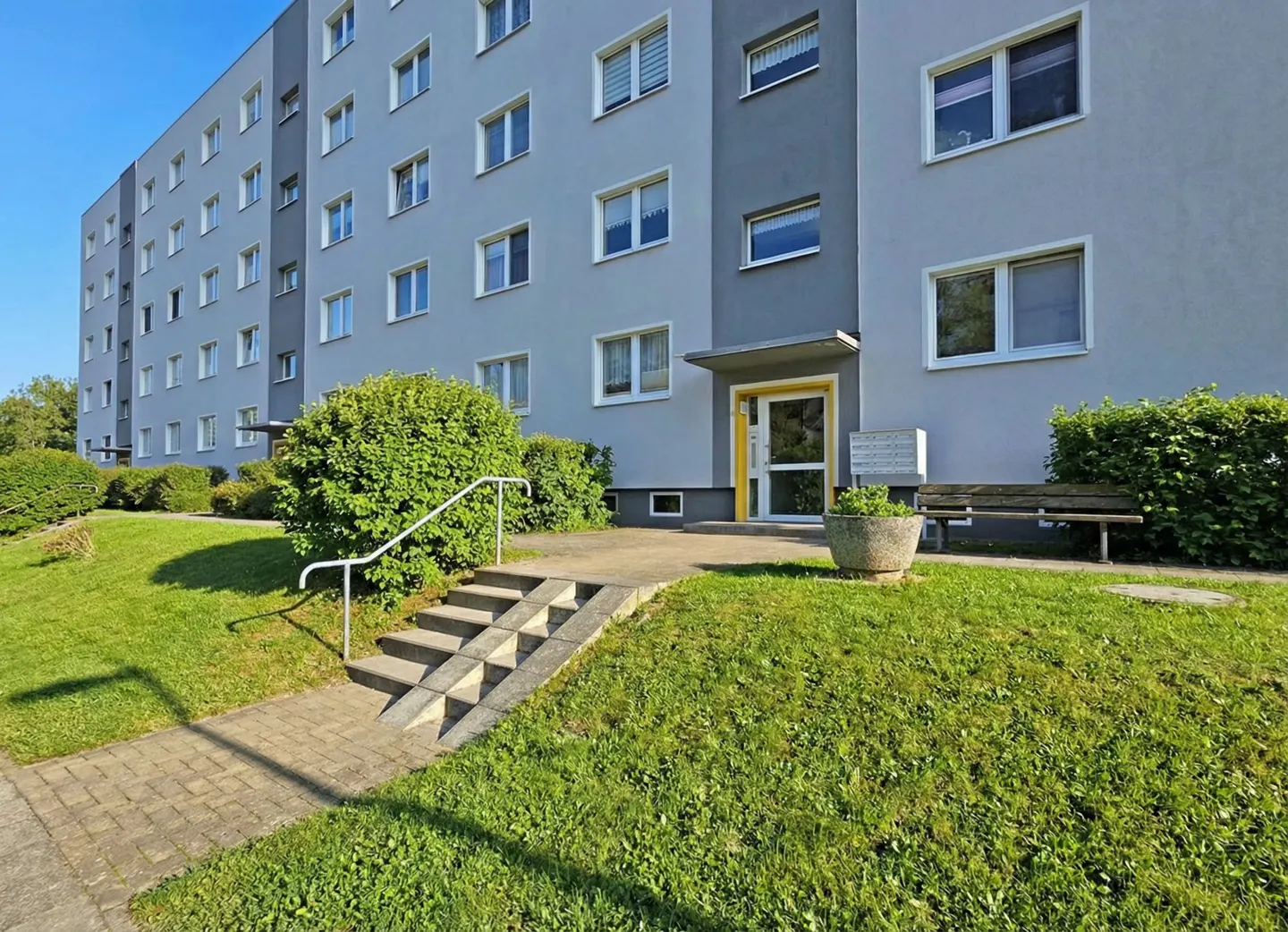 Exterior view of a grey apartment building with a yellow-framed entrance, concrete steps, and a green lawn with bushes.