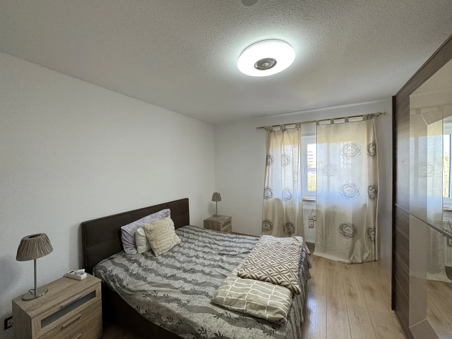Bedroom with a queen bed, wood floors, white walls, and beige curtains. Nightstands with lamps flank the bed. A large wardrobe stands to the right.