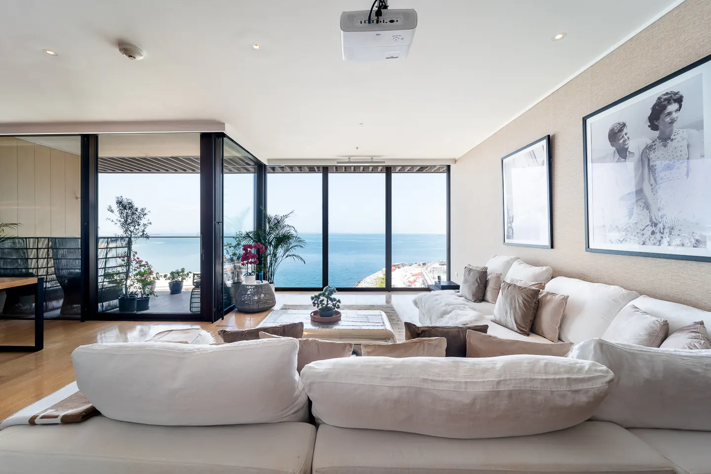 Bright living room with a large white sofa, pillows, and ocean view through floor-to-ceiling windows. A projector hangs from the ceiling.