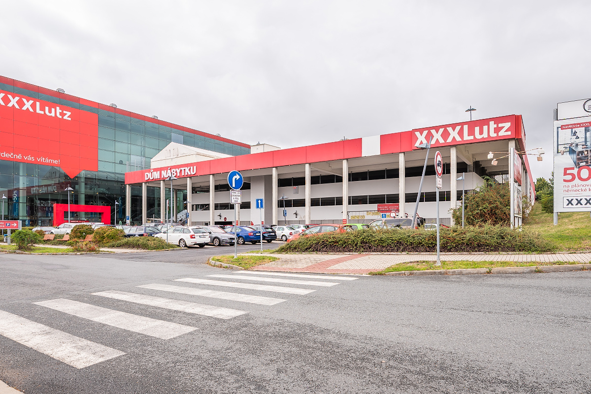 Exterior view of a large XXXLutz furniture store with a parking lot, crosswalk, and red and white branding on a cloudy day. [AI-generated text]
