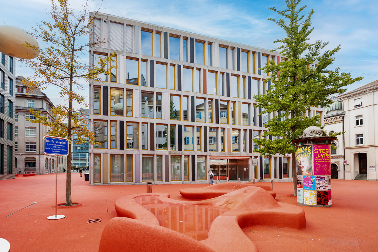 Modern office building with a grid facade, set in a public square with bright red ground, trees, and a sculptural fountain. [AI-generated text]