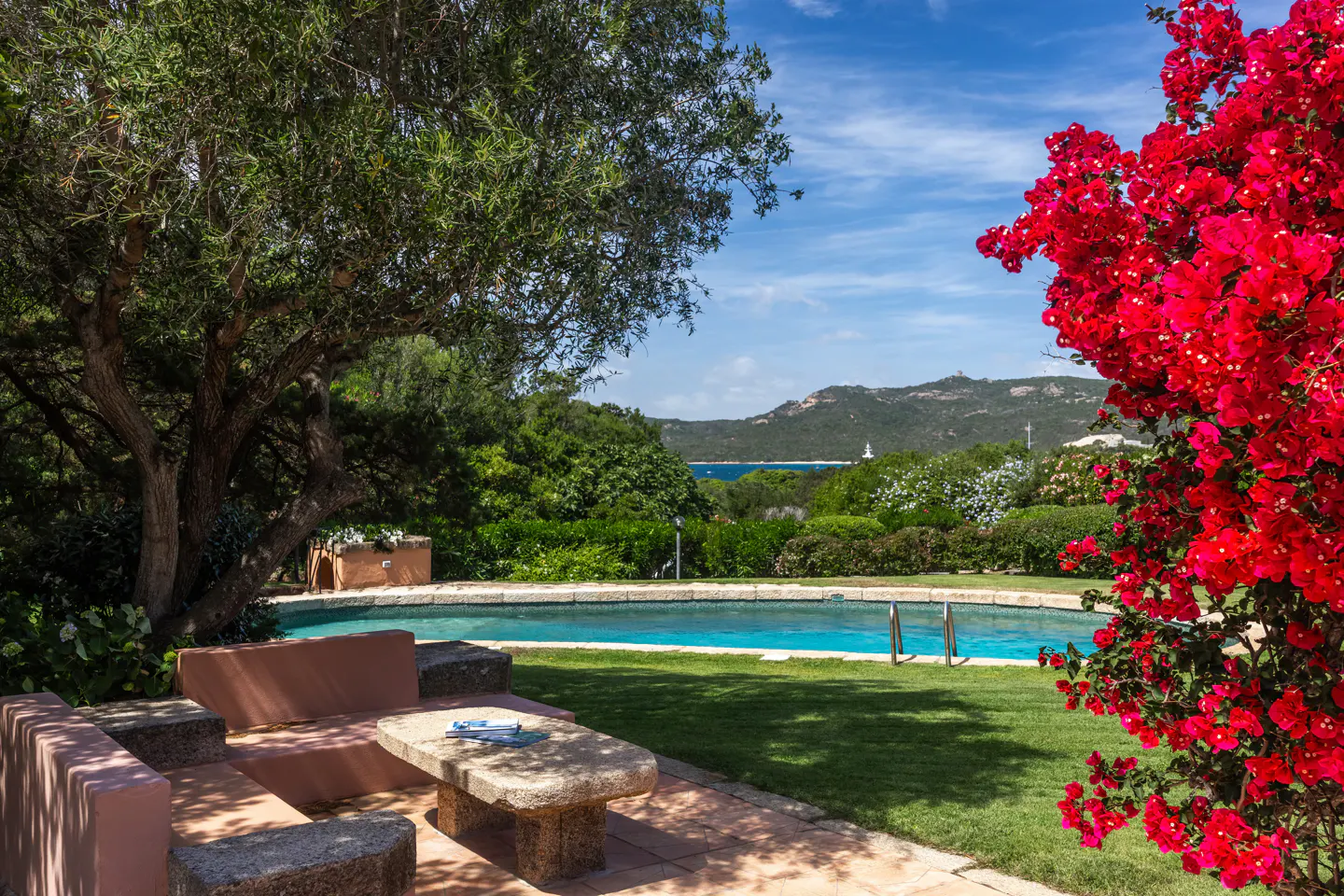 A stone patio with a table and bench overlooks a swimming pool, lush greenery, and a distant mountain view under blue sky.