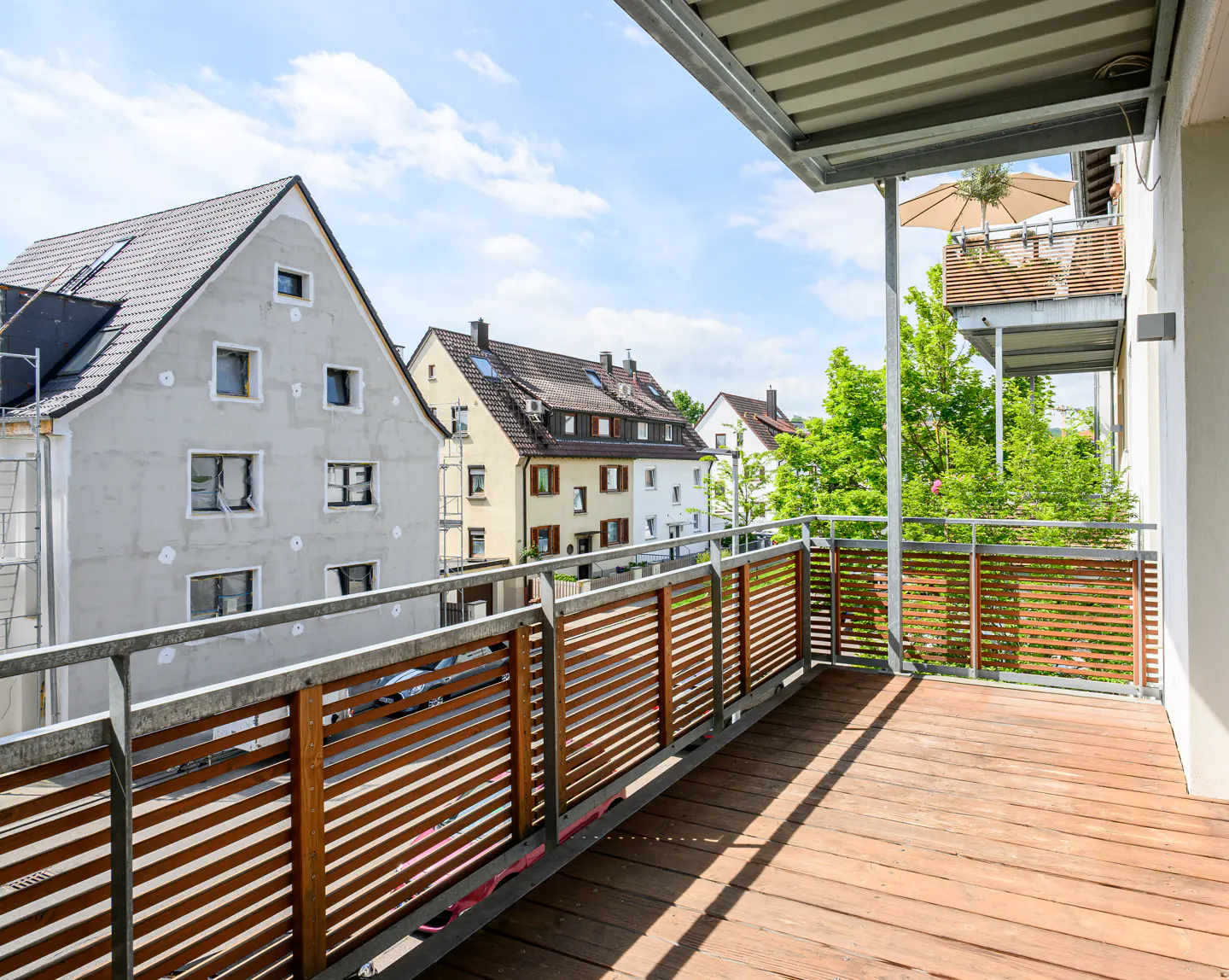 A wood-floored balcony with a metal railing overlooks houses and green trees under a blue sky.