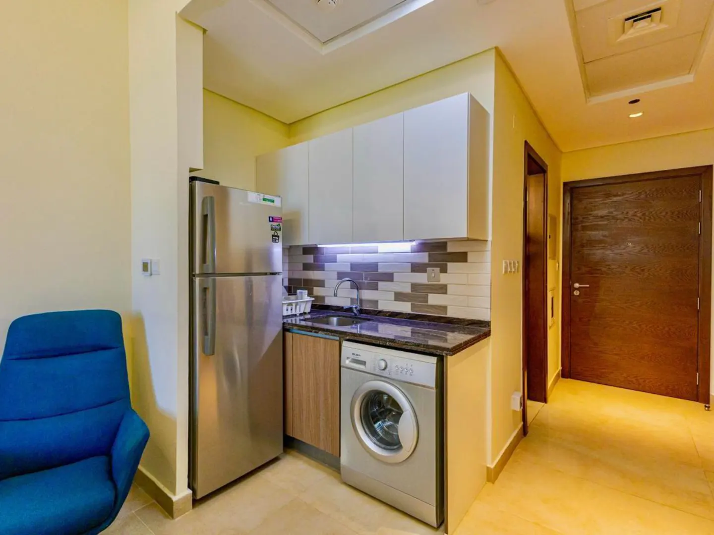 A compact kitchen area with a stainless steel fridge, washer, sink, and white cabinets. A blue chair sits nearby.