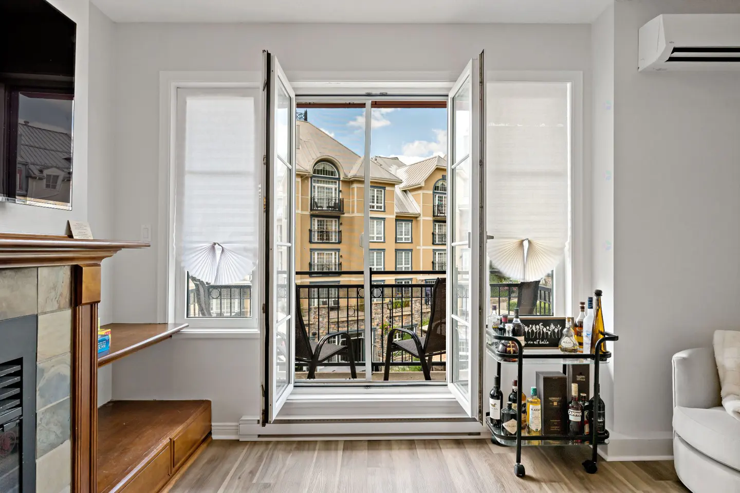 Living room with open French doors to a balcony with chairs, overlooking a building. A bar cart with bottles is on the right.