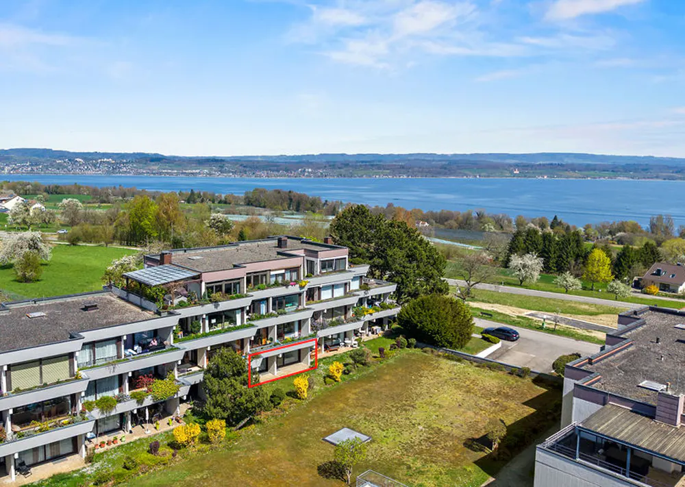 Aerial view of a modern apartment building with a red box highlighting a unit, overlooking a lake and green landscape.