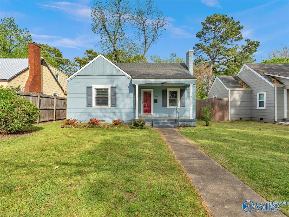 A light blue house with a red door and gray shutters sits on a green lawn under a blue sky.