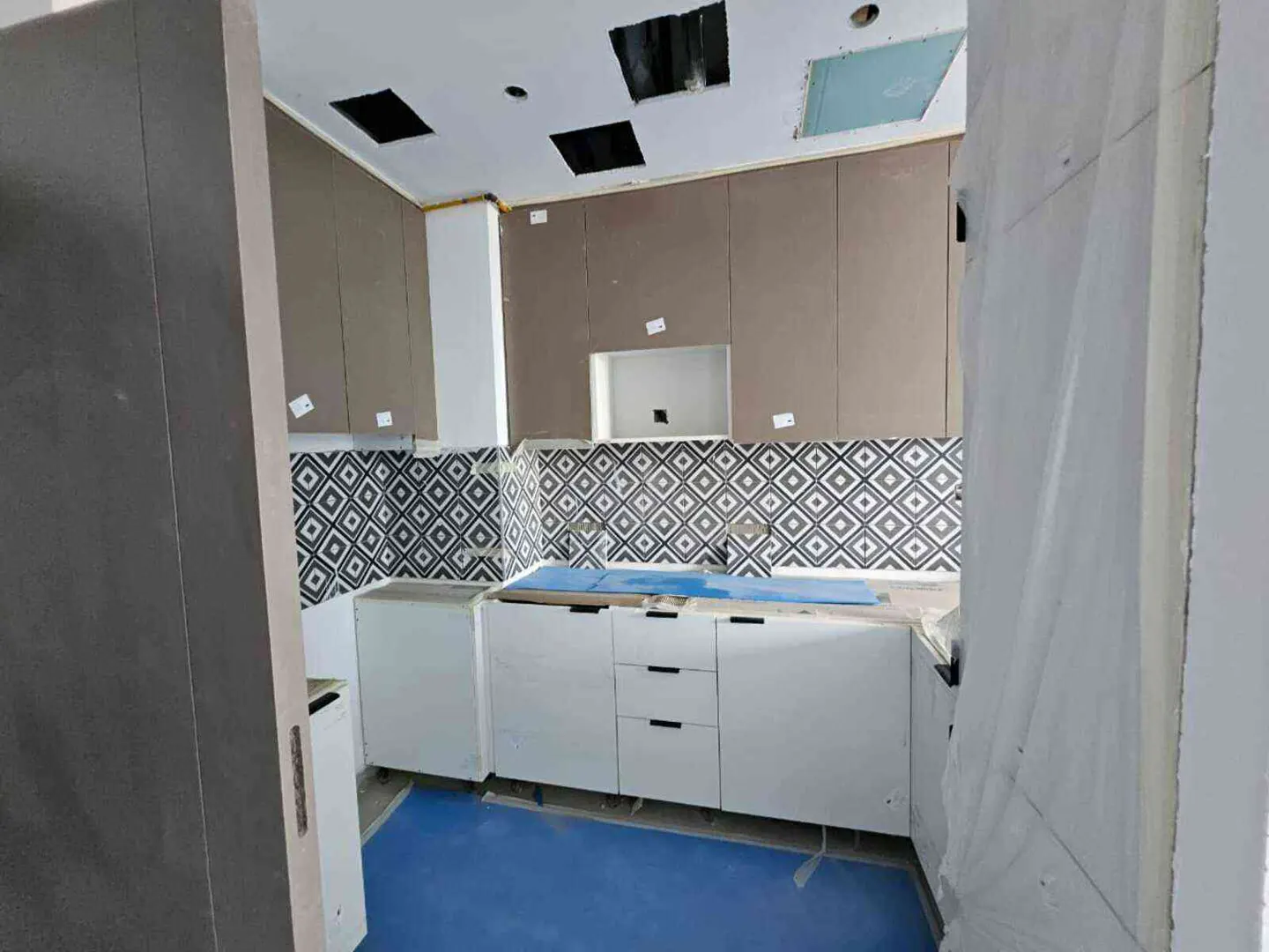 A kitchen under construction with white cabinets, brown upper cabinets, and patterned backsplash. Blue floor covering.