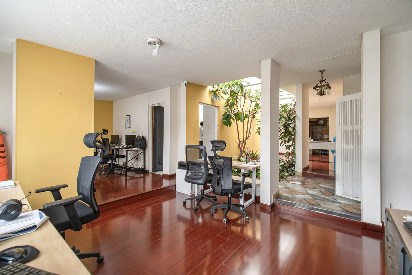 Office space with hardwood floors, yellow walls, and black office chairs. A courtyard with plants is visible through an opening.