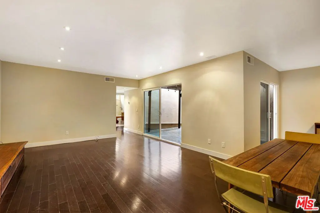 A bright, empty room with dark wood floors, beige walls, and recessed lighting. A sliding glass door leads to an outdoor space. A wooden table and chairs are in the foreground.