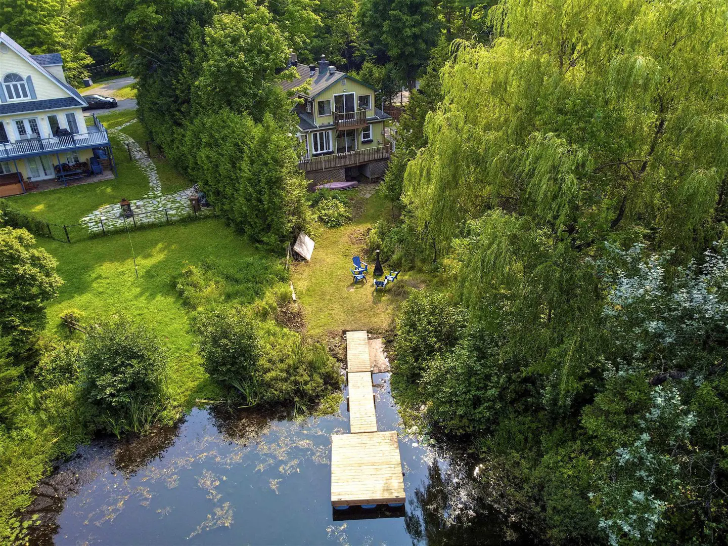 Aerial view of a lake house with a wooden dock leading to the water, surrounded by lush green trees.