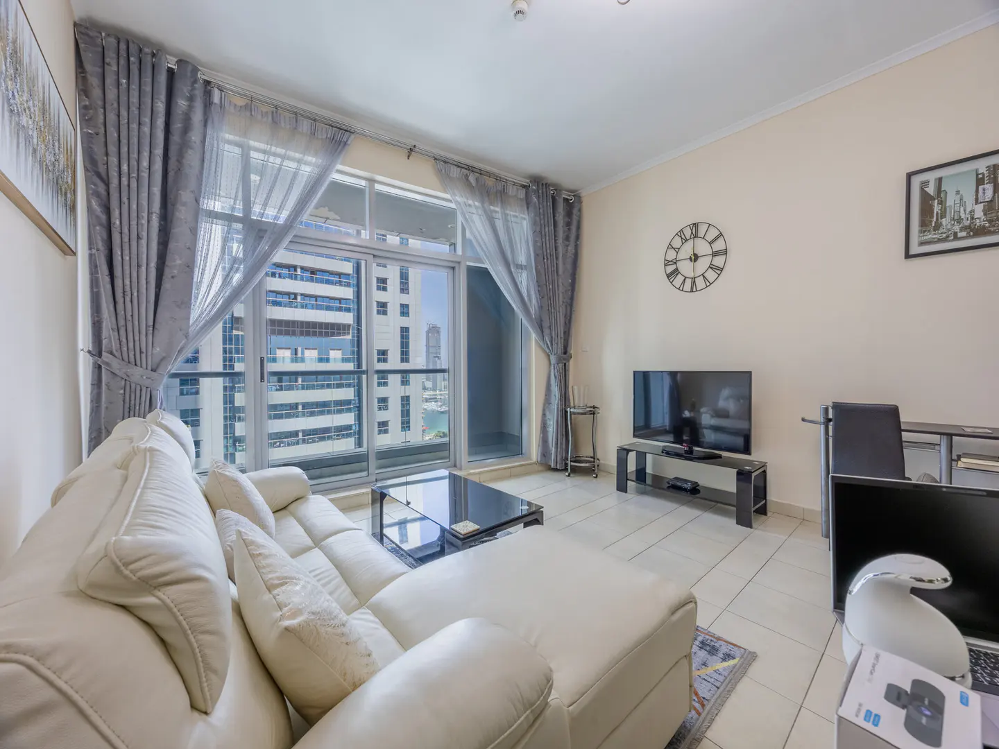Living room with white leather sofa, black glass coffee table, TV, and large window with city view. Gray curtains frame the window.