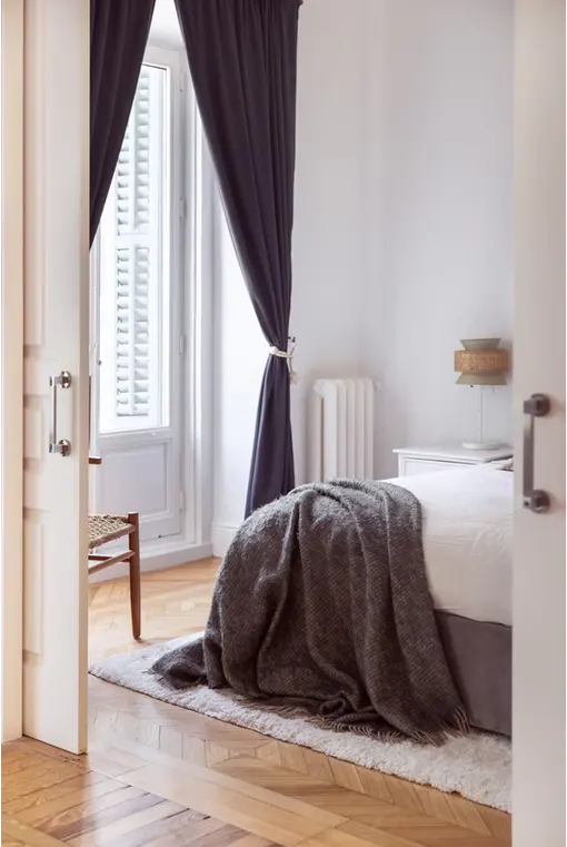 Bedroom with white walls, hardwood floors, and a bed with a gray blanket. Gray curtains frame a window with white shutters.