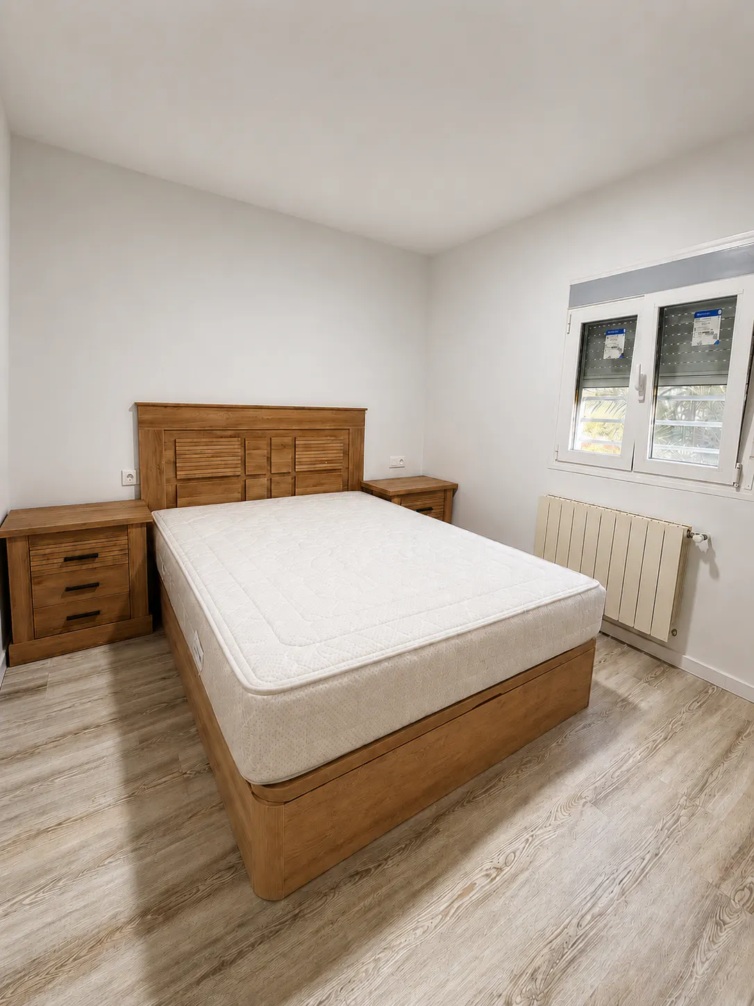 A bedroom with a wooden bed frame, white mattress, matching nightstands, light wood floors, and a window with a radiator.