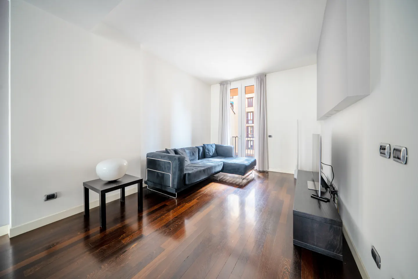 Bright living room with dark wood floors, white walls, and a blue velvet sectional sofa. A black table with a white vase sits near a window.