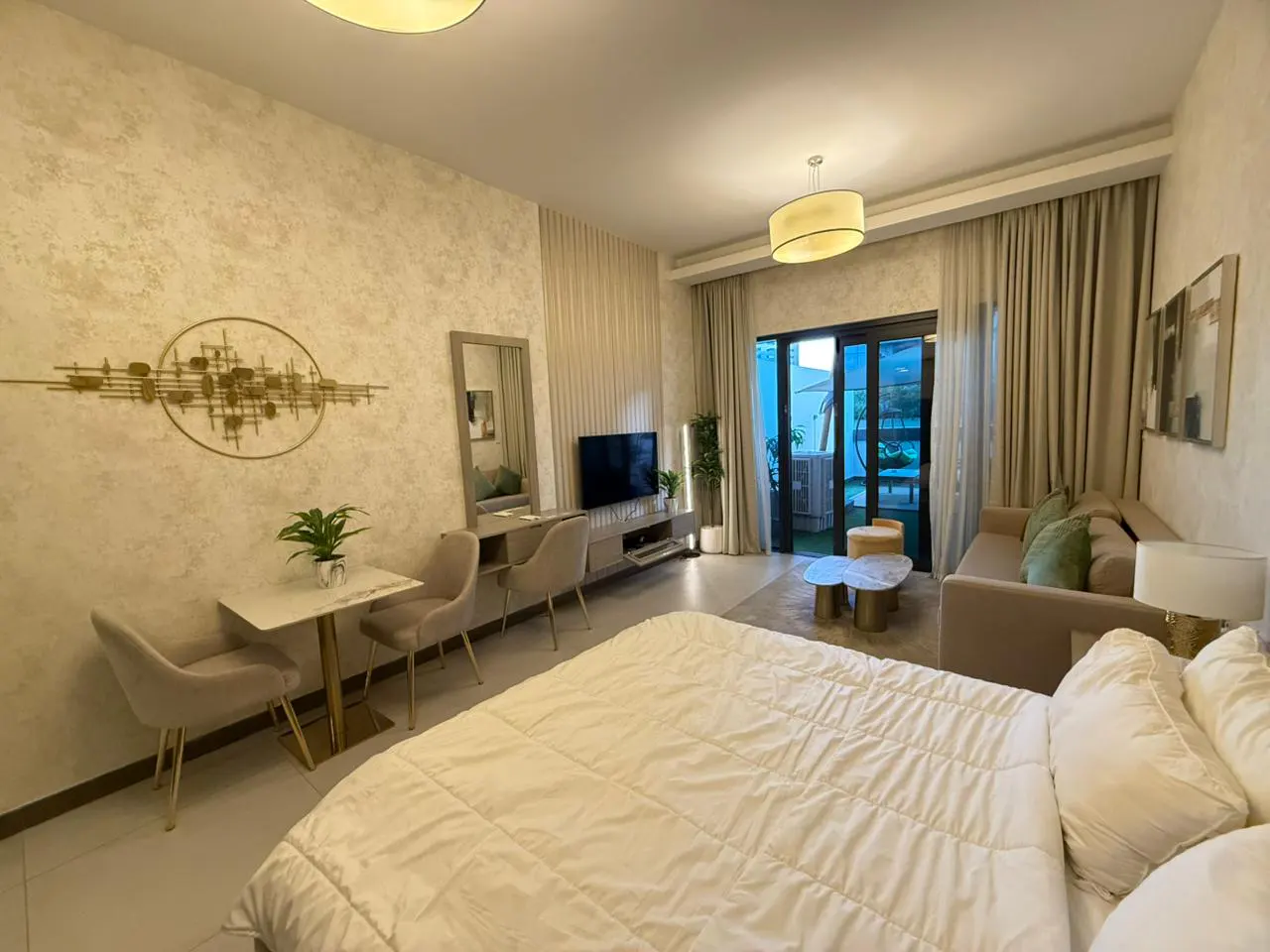 A modern studio apartment with a white bed, dining table, TV, and sofa. Neutral tones and natural light create a cozy atmosphere.