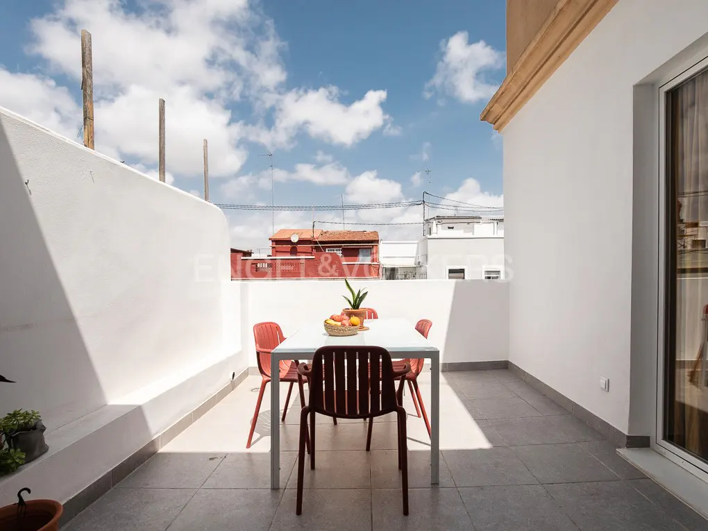 A bright, sunny patio with a white table and red chairs. A basket of fruit sits on the table. White walls surround the patio.