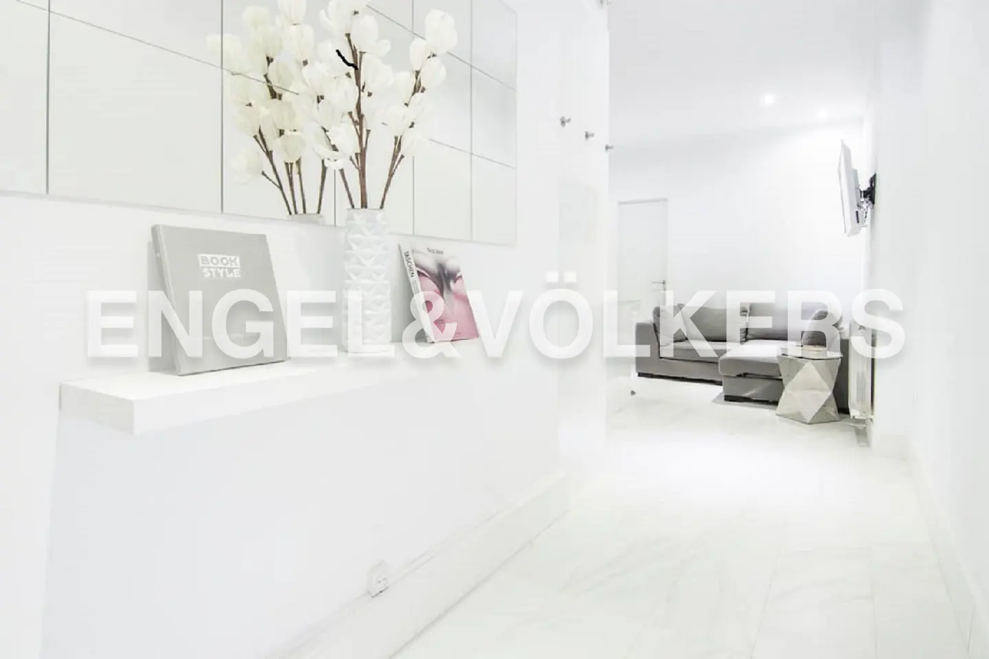 Bright white hallway with a shelf holding a book, vase with white flowers, and a picture. A gray couch is visible in the background.
