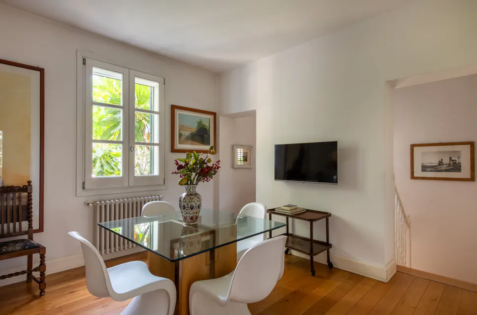 Dining area with a glass table, four white modern chairs, a floral vase, wall-mounted TV, and hardwood floors.