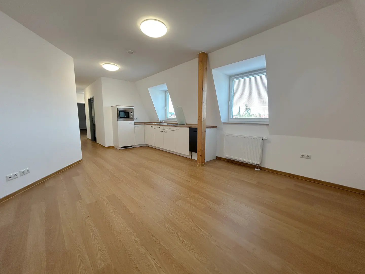 Bright, empty apartment with wood floors, white walls, and a small kitchen area with white cabinets and a microwave. Two windows let in natural light.