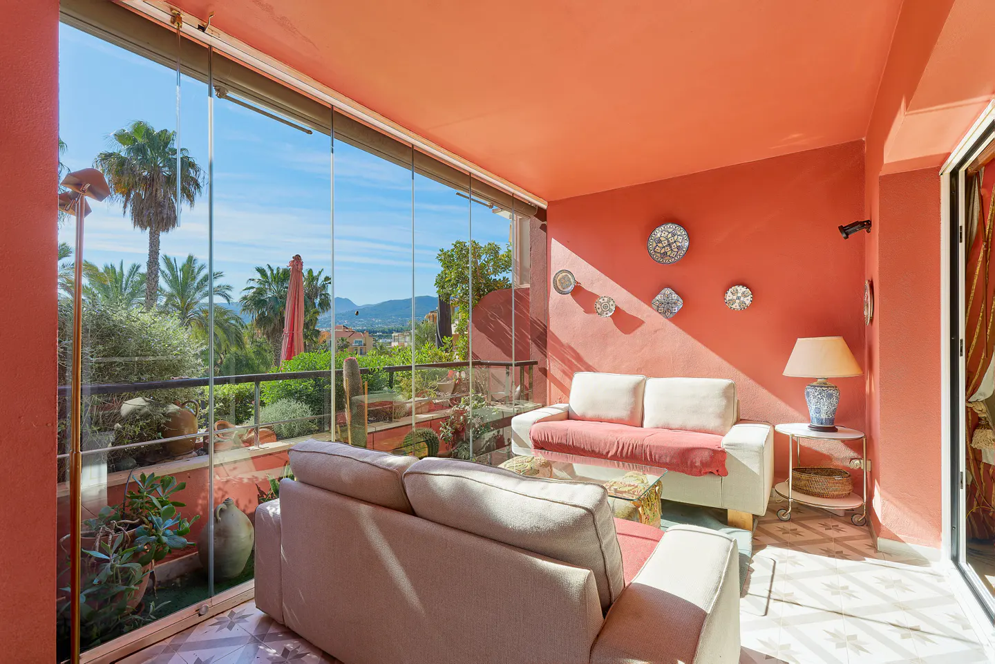 A sunroom with coral walls, tile floors, and glass doors opening to a garden view. Two sofas, a lamp, and wall decorations furnish the space.
