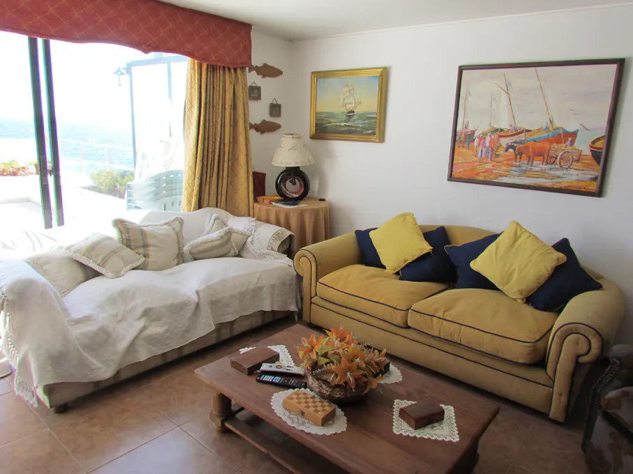 Living room with a white sofa, a yellow sofa with blue and yellow pillows, and a wooden coffee table. Paintings on the wall.