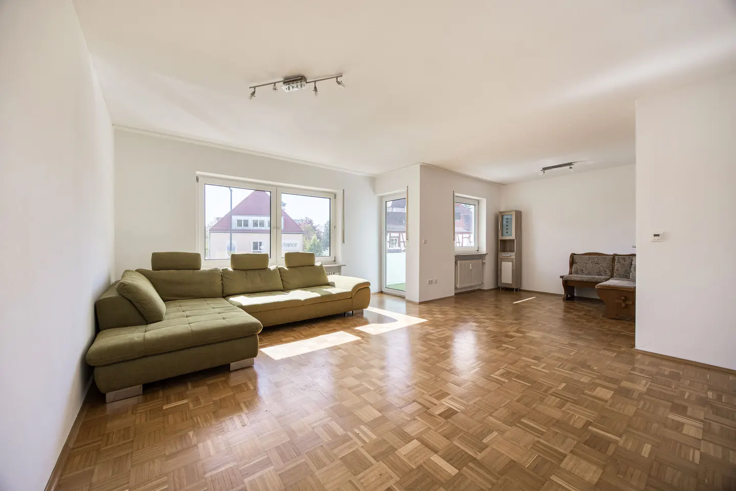 Bright living room with parquet floors, white walls, and a green sectional sofa by a large window.