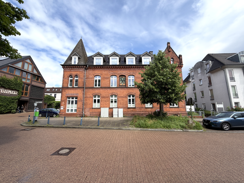 A historic red brick building with a turret and dormer windows stands beside a tree, viewed from a paved town square.