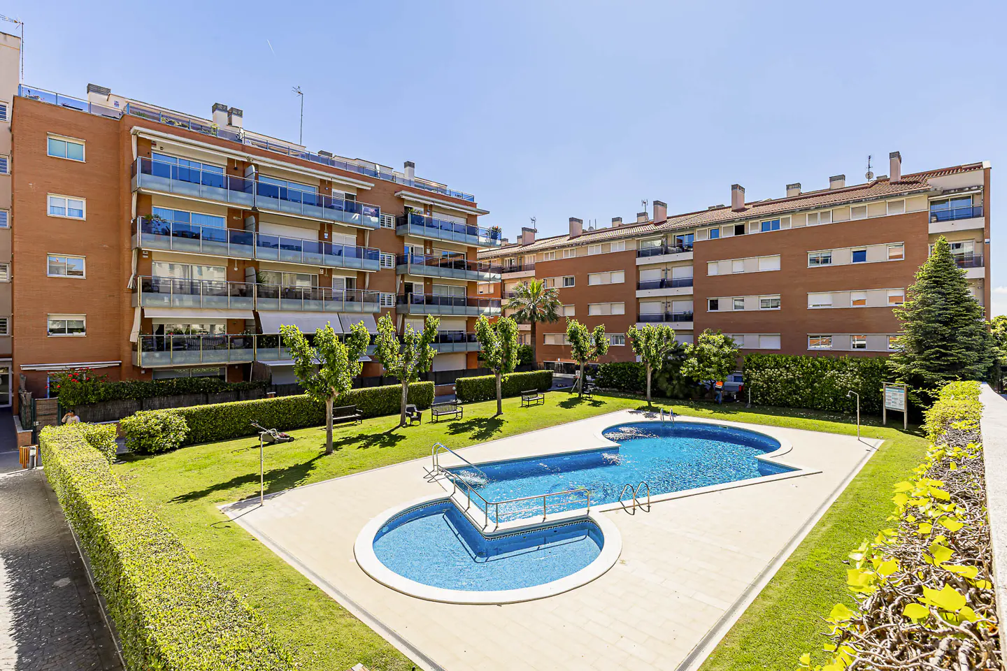 Apartment complex with a blue swimming pool, green grass, trees, and brick buildings under a clear blue sky.