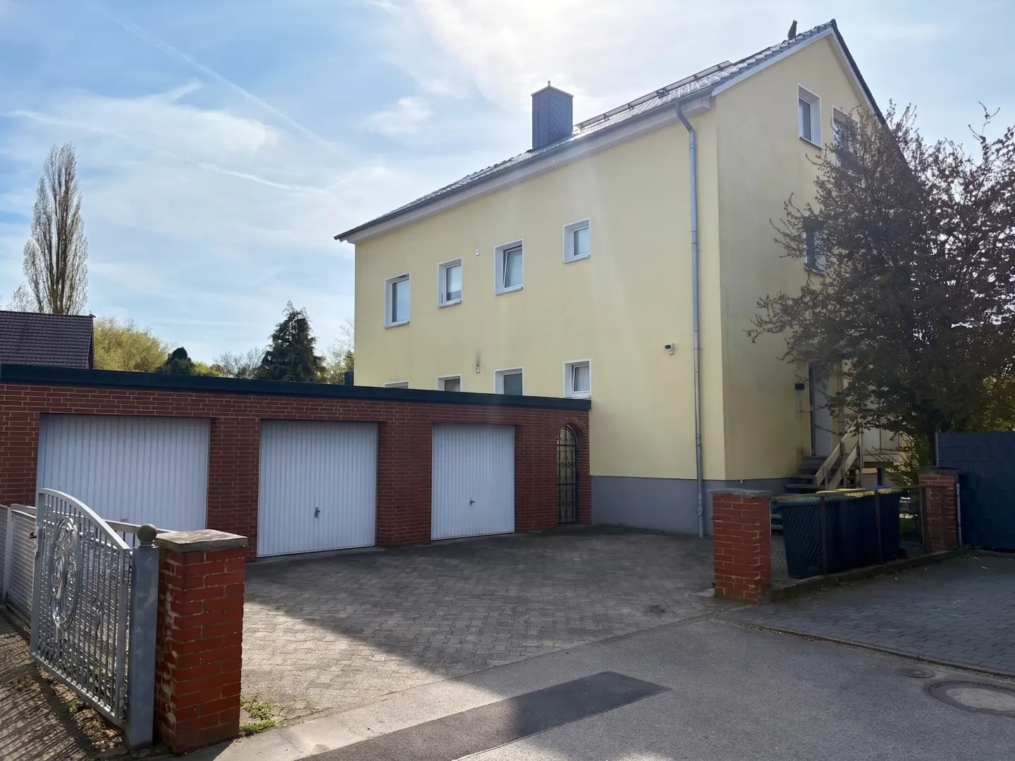 Two-story yellow house with a three-car garage. The garage has red brick walls and white doors. A gray metal gate is on the left.