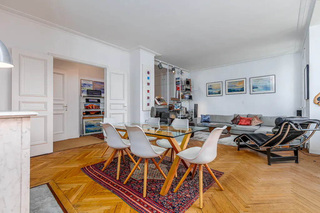 Bright living room with parquet floors, white walls, and a glass dining table with white chairs on a patterned rug. A gray sofa and black lounge chair are in the background.