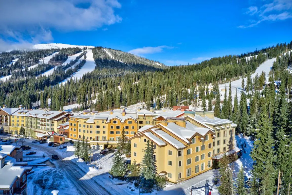 Scenic view of a ski resort with yellow buildings, snow-covered mountains, and evergreen trees under a bright blue sky.