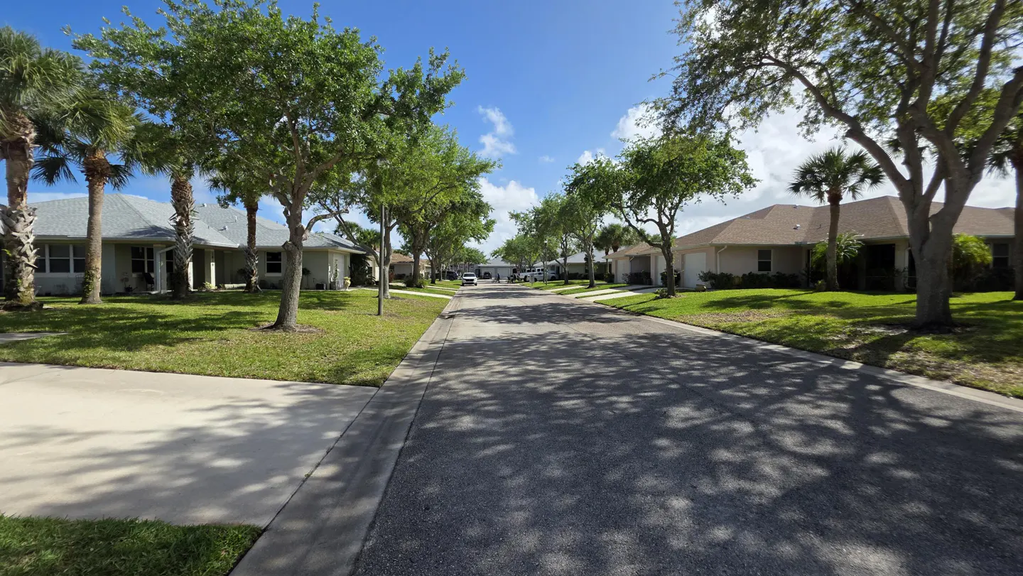 Residential street view with houses, green lawns, and trees lining the road under a blue sky with scattered clouds.