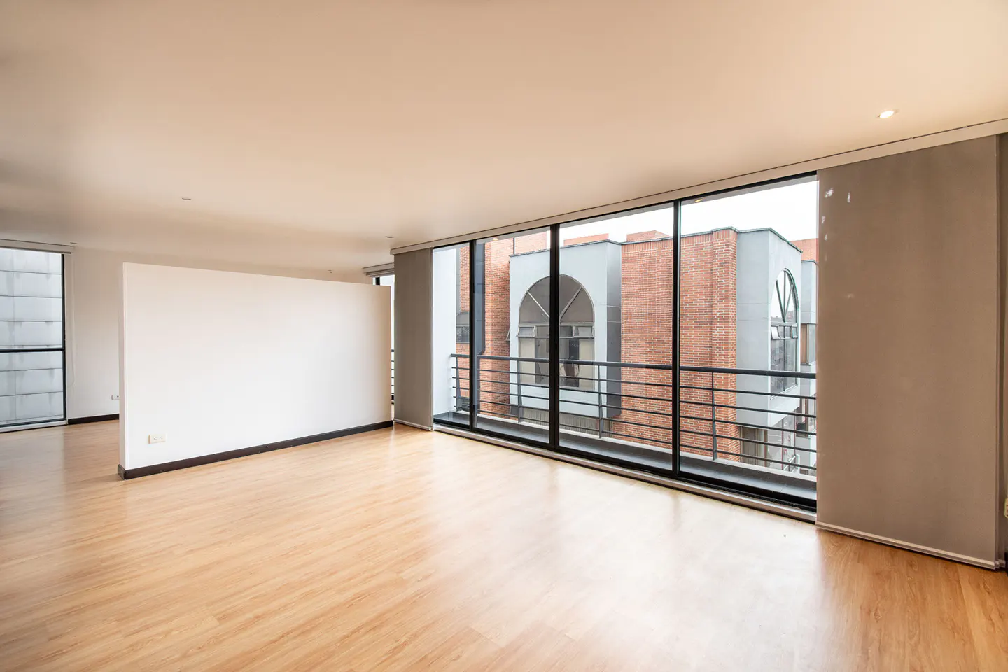 Empty room with wood floors, white walls, and large windows. Outside view of brick building with arched windows and black railings.